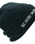 Black beanie with 'BE HERE NOW' text on a white background