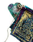 A close-up of a multicolored fabric pouch or bookmark with a rustic twine loop, featuring a prominent gold foil stamp design on a dark blue background.