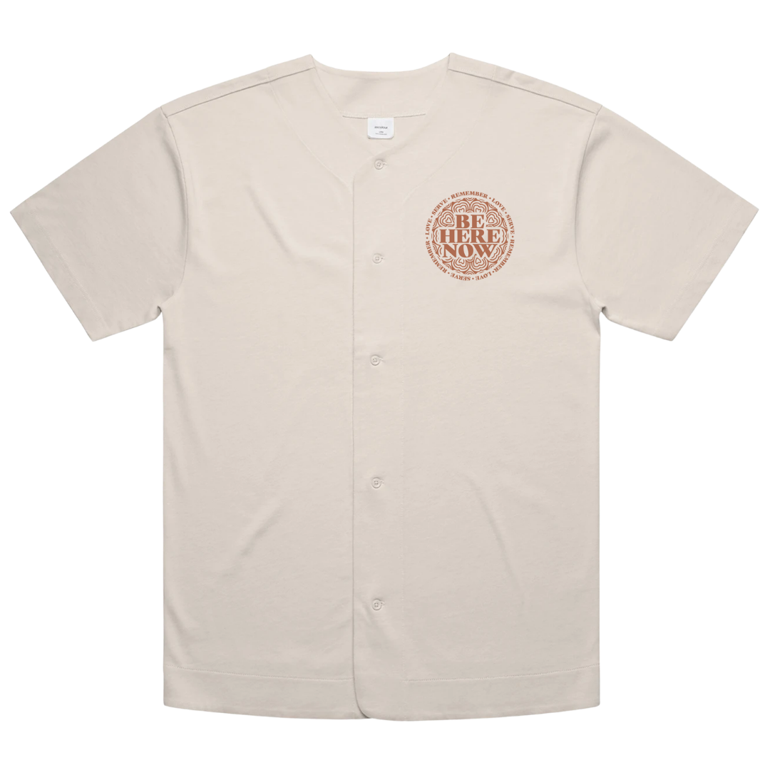 A short-sleeve button-front baseball-style shirt in a light warm beige color, featuring a circular chest graphic with the words 'BE HERE NOW' in a textured reddish-brown font. The shirt is displayed flat against a solid white background.