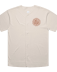 A short-sleeve button-front baseball-style shirt in a light warm beige color, featuring a circular chest graphic with the words 'BE HERE NOW' in a textured reddish-brown font. The shirt is displayed flat against a solid white background.