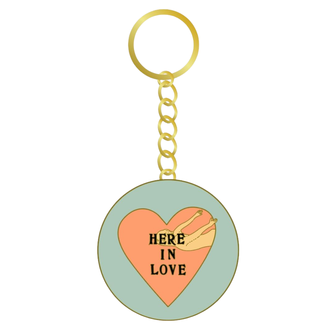 A round enamel-style keychain charm with a gold-tone split ring and chain. The charm features a pale mint green background with a large peach heart in the center, a small beige bird illustration, and the words “HERE IN LOVE” in black text.