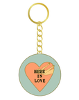 A round enamel-style keychain charm with a gold-tone split ring and chain. The charm features a pale mint green background with a large peach heart in the center, a small beige bird illustration, and the words “HERE IN LOVE” in black text.