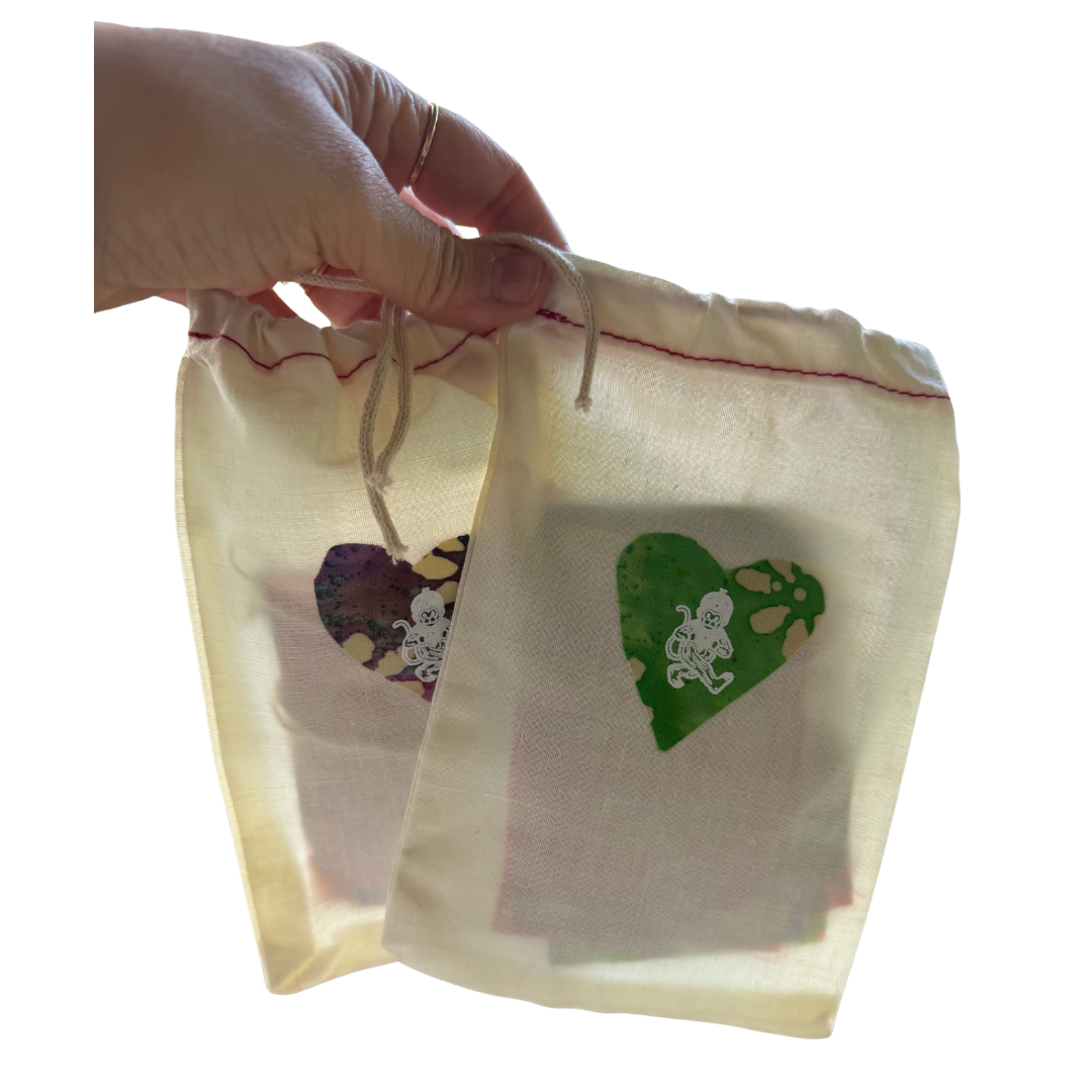 A hand holds two small cream-colored drawstring pouches with printed heart designs. The pouches appear semi-transparent, showing a light background through the fabric, with one heart in green and the other in a multicolor floral pattern.