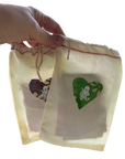 A hand holds two small cream-colored drawstring pouches with printed heart designs. The pouches appear semi-transparent, showing a light background through the fabric, with one heart in green and the other in a multicolor floral pattern.