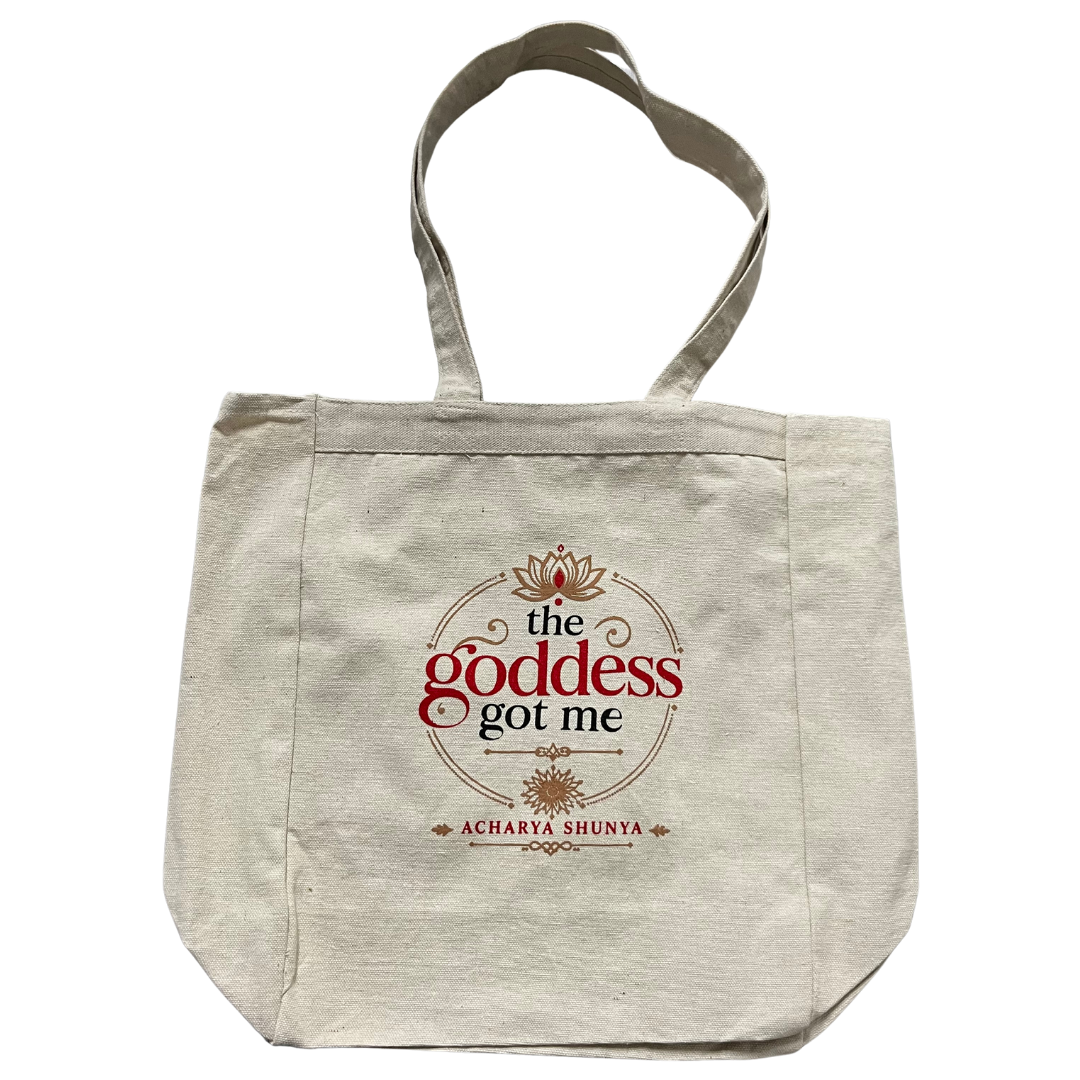 A natural canvas tote bag with a printed graphic on the front that reads 'the goddess got me' and 'ACHARYA SHUNYA' in red and black text with decorative gold details.