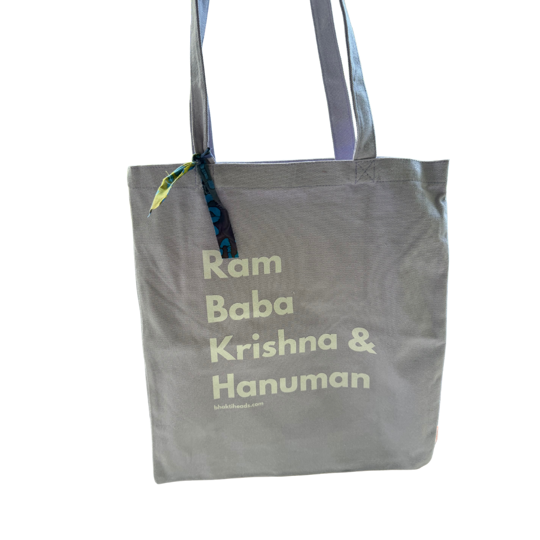 A gray canvas tote bag with the text 'Ram Baba Krishna &amp; Hanuman' printed in a light cream font. The bag has gray shoulder straps and a small green and blue patterned ribbon tied to the top left corner.