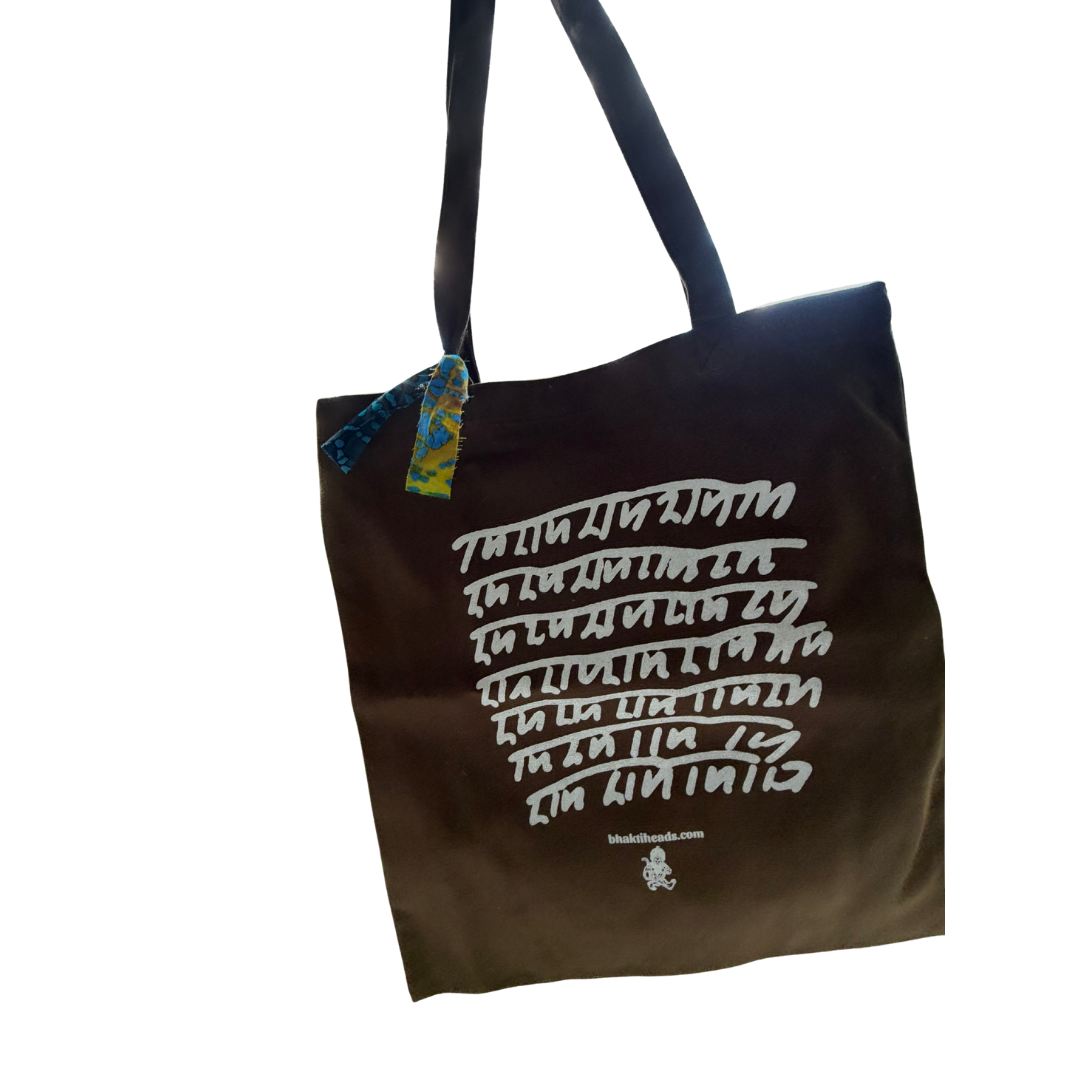 A dark brown canvas tote bag with black shoulder straps, featuring white Ram Ram script printed on the front. A small blue and yellow patterned tag is attached near the top left corner, and a small logo is printed near the bottom.