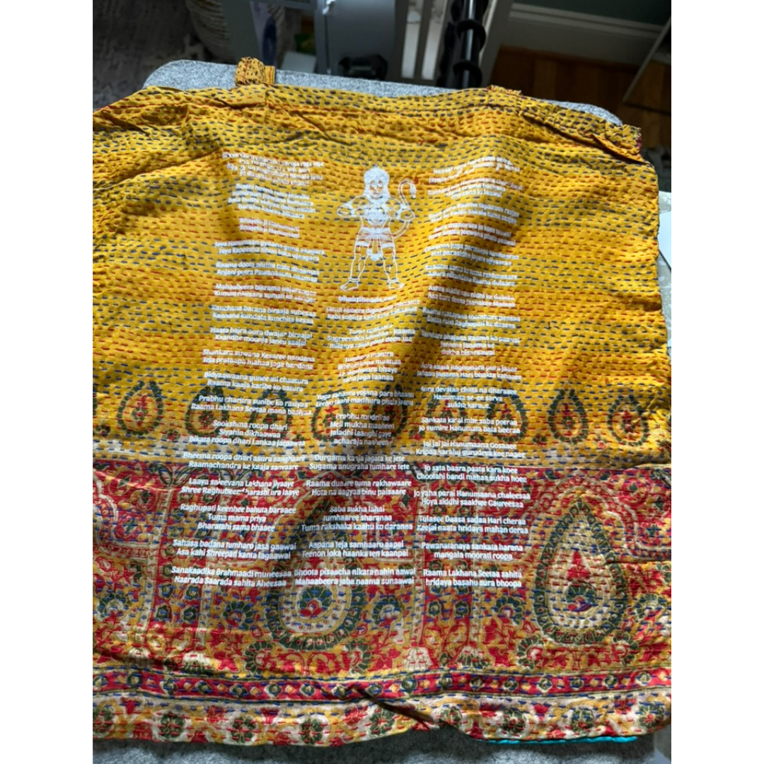 A yellow and red patterned fabric garment, likely a scarf or shawl, laid flat on a gray surface. The top portion is a textured mustard yellow with white text and a central white figure, while the bottom portion features a detailed red, orange, and green paisley and floral pattern.