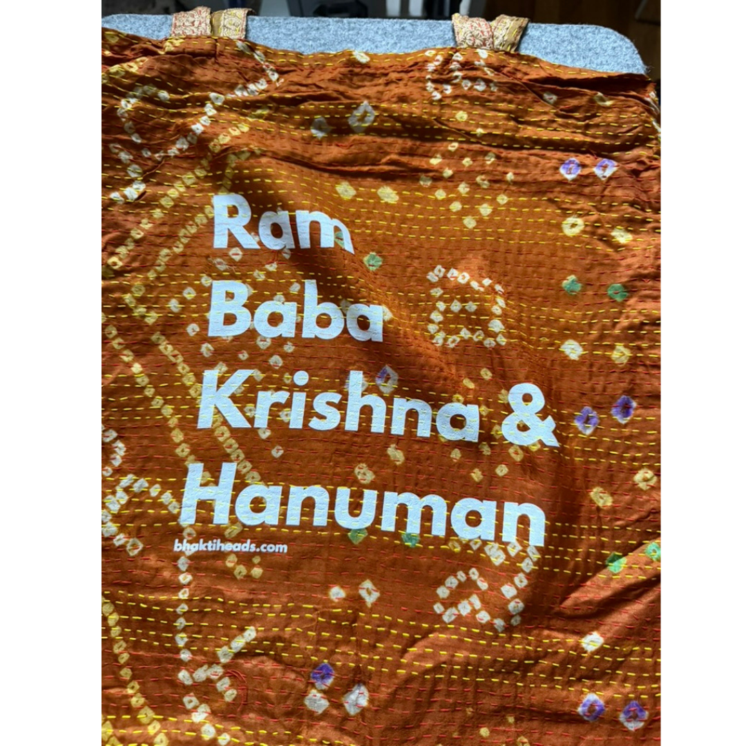 A brown and orange patterned fabric cloth with white text reading 'Ram Baba Krishna &amp; Hanuman' and a small website watermark, photographed flat against a dark background.