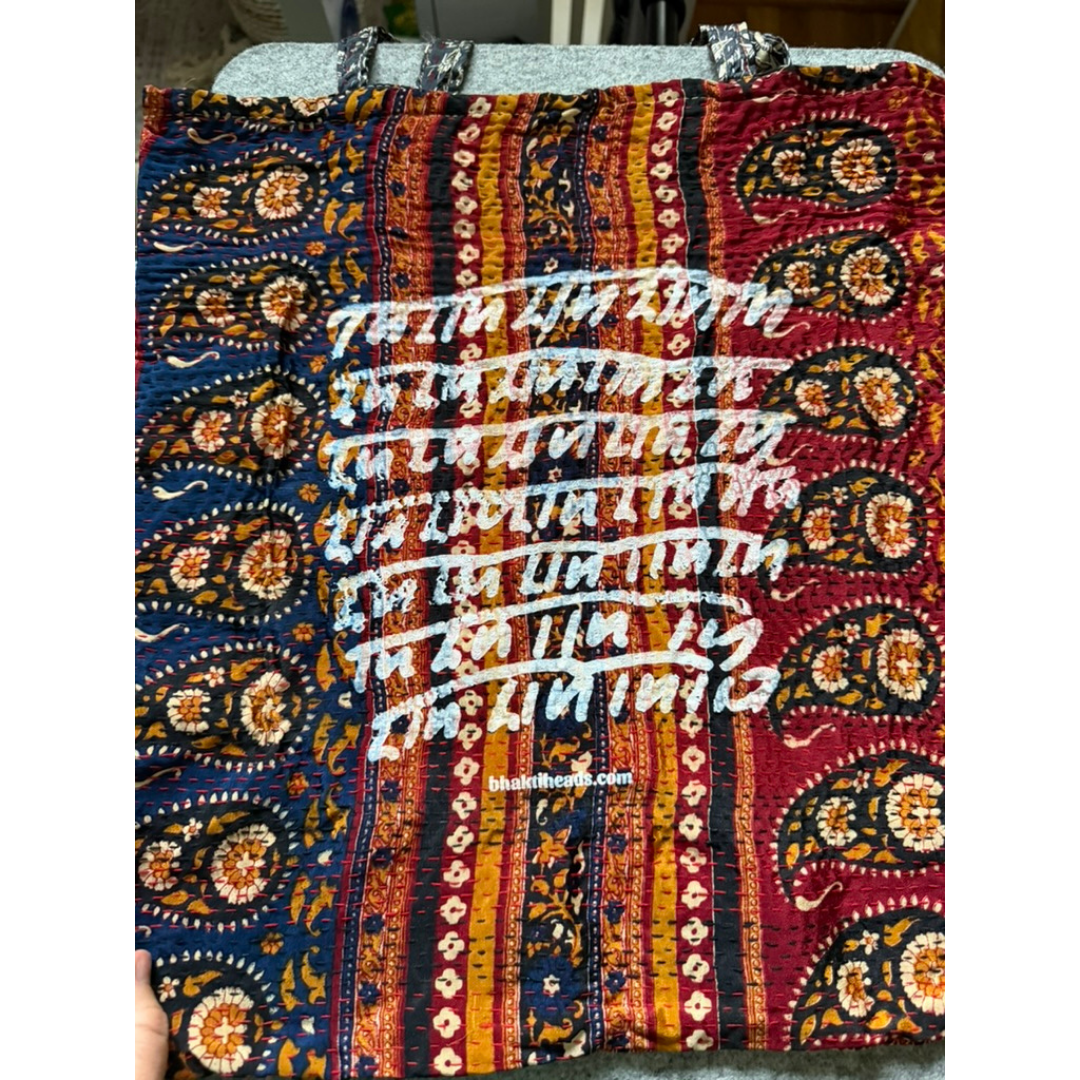 A folded patterned textile with vertical multicolor stripes and paisley motifs in red, blue, orange, yellow, and black, overlaid with repeated white script text. A small visible watermark reads 'TheHandmadeShop.