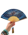 A hand holds a folding hand fan with a blue illustrated design depicting a figure on a wave, with a yellow-gold handle. A matching card tag hangs from the handle, and the background is solid white.